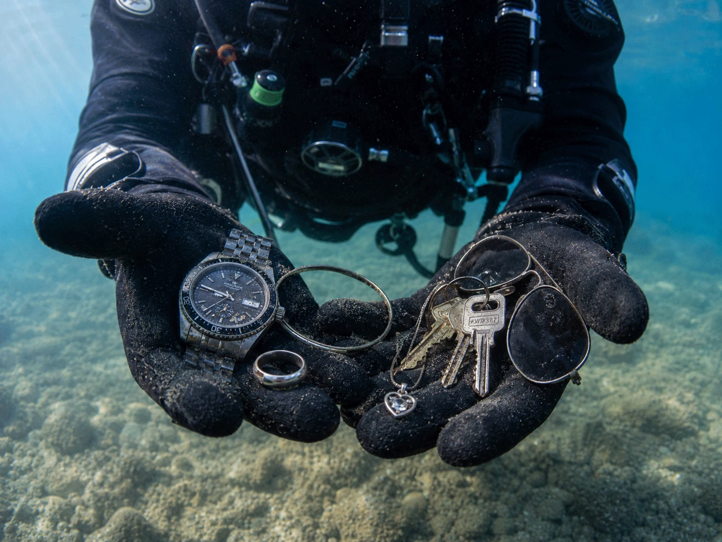 A diver holding recovered personal items including a watch, ring, keys, glasses, and jewelry
